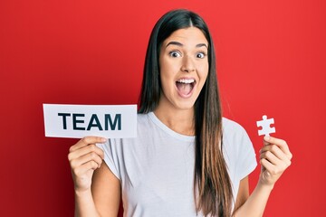 Poster - Young brunette woman holding team paper and piece of puzzle as teamwork celebrating crazy and amazed for success with open eyes screaming excited.