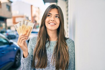 Young beautiful hispanic girl smiling happy holding norwegian krone banknotes at the city.