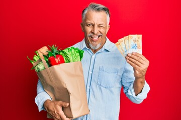 Sticker - Middle age grey-haired man holding groceries and swiss franc banknotes winking looking at the camera with sexy expression, cheerful and happy face.