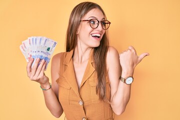 Young blonde girl holding colombian pesos pointing thumb up to the side smiling happy with open mouth