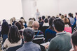 © kasto - Business and entrepreneurship symposium. Female speaker giving a talk at business meeting. Audience in conference hall. Rear view of unrecognized participants making notes in audience.