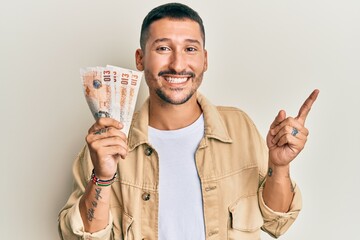 Canvas Print - Handsome man with tattoos holding 10 united kingdom pounds banknotes smiling happy pointing with hand and finger to the side