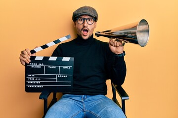 Poster - Handsome man with tattoos holding video film clapboard and louder afraid and shocked with surprise and amazed expression, fear and excited face.