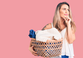 Poster - Young beautiful blonde woman holding laundry basket serious face thinking about question with hand on chin, thoughtful about confusing idea