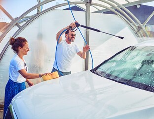 Wall Mural - Middle age beautiful couple wearing casual clothes and smiling happy. Standing with smile on face washing car using water pistol and sponge.