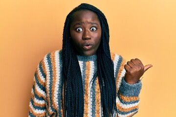 Sticker - Young black woman with braids wearing casual winter sweater surprised pointing with hand finger to the side, open mouth amazed expression.