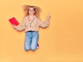 Young beautiful curly student girl wearing glasses smiling happy. Jumping with smile on face reading book over isolated yellow background.