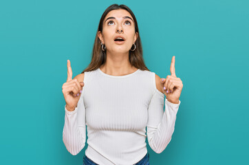 Young hispanic woman wearing casual clothes amazed and surprised looking up and pointing with fingers and raised arms.