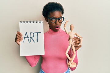 Poster - Young african american girl holding wooden manikin and art word on notebook in shock face, looking skeptical and sarcastic, surprised with open mouth