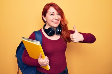 Poster - Young beautiful redhead student woman wearing backpack and headphones holding notebook smiling happy and positive, thumb up doing excellent and approval sign