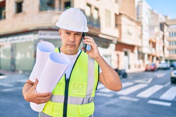 Middle age architect man talking on the smartphone and holding blueprints at the city.