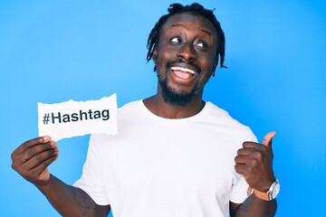 Poster - Young african american man with braids holding hashtag paper pointing thumb up to the side smiling happy with open mouth