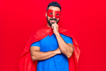 Sticker - Young hispanic man wearing super hero costume smiling looking confident at the camera with crossed arms and hand on chin. thinking positive.