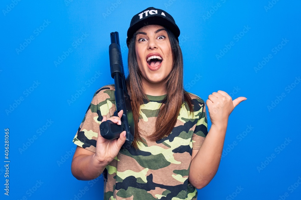 Young beautiful hunter woman wearing camouflage t-shirt and usa cap holding shotgun pointing thumb up to the side smiling happy with open mouth
