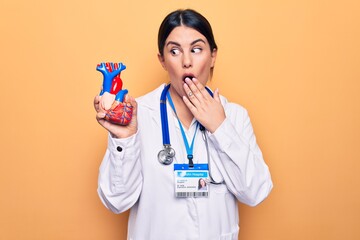 Poster - Young beautiful cardiologist woman wearing stethoscope holding heart over yellow background covering mouth with hand, shocked and afraid for mistake. Surprised expression