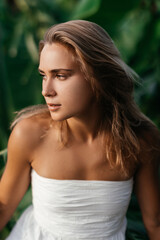 Fashion outdoor portrait of blonde woman, wearing elegant white dress, tropical leaves on background.