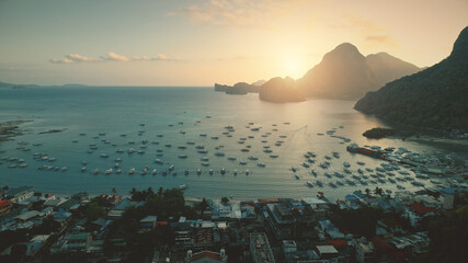 Wall Mural - Aerial sunset at ocean harbor with boats of tropic port town cityscape. Buildings, lodges, homes with resort at sand coast. Mount Philippines island silhouette at sun light. Cinematic drone shot