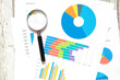 © Alena - The magnifying glass lies on papers with color charts on white wooden background. Analyzing finances. Graph. Top view. Business concept.