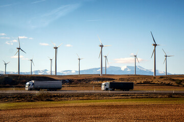 Dos camiones que transportan mercancía por carretera circulando en la autovía con los molinos eólicos de energía limpia y las montañas nevadas de fondo .