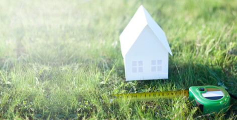 Miniature paper house on green grass in summer. Mortgage. Measuring tape or tape measure. The concept of buying a land plot for building a house.