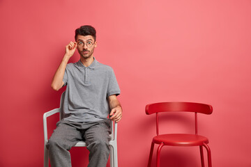 Poster - Surprised young man with stubble stares through spectacles poses on chair hears astonishing news dressed in t shirt and trousers isolated over coral background. People shock reaction concept.