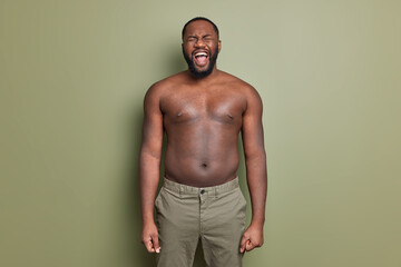 Wall Mural - Emotional dark skinned man with naked torso wears shorts screams loudly opens mouth widely has thick beard poses against khaki color background keeps arms down. Screaming guy. Emotions concept