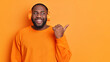 © WHstudio Leushin N - Studio shot of joyful bearded black man listens audio track via wireless headphones wears bright orange long sleeved jumper in one tone with background points thumb away advertises copy space