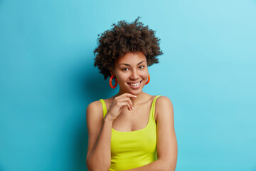 Wall Mural - portrait of pleasant looking curly woman keeps hand under chin smiles gently feels satisfied dressed