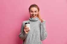 Overjoyed Millennial European Girl Has Coffee Break Holds Paper Cup Of Coffee Raises Hand Hears Great News Wears Big Optical Glasses And Grey Jumper Poses With Beverage Against Pink Background