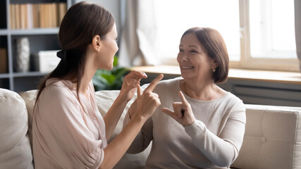 Wall Mural - Close up smiling mature mother and grownup daughter speaking sign language, sitting on couch, young woman with elderly mum enjoying pleasant conversation, communicating, showing gestures, deaf family