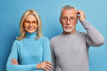 Wall Mural - Studio shot of smiling blonde European woman keeps arms folded looks directly at camera. Puzzled mature man scratches head and stares in bewilderment. Portrait of family couple. Blue background