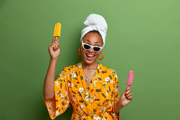 Wall Mural - Positive dark skinned young woman dressed in domestic clothes wears trendy sunglasses bath towel on head and dressing gown holds delicious ice cream poses against green background. Summer dessert