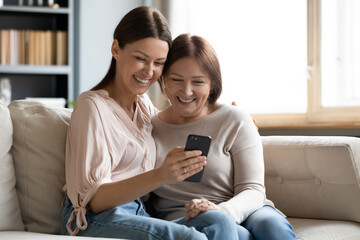 Wall Mural - Close up happy mature mother and grownup daughter using phone together, hugging, sitting on cozy couch at home, smiling young woman embracing elderly mum, watching funny video, shopping online