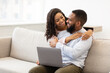© Prostock-studio - African american couple sitting on couch, using laptop, cuddling
