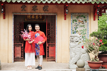 Sticker - Cheerful young Vietnamese couple in traiditional dreses standing at temple doors with blooming peach branches and paper fan