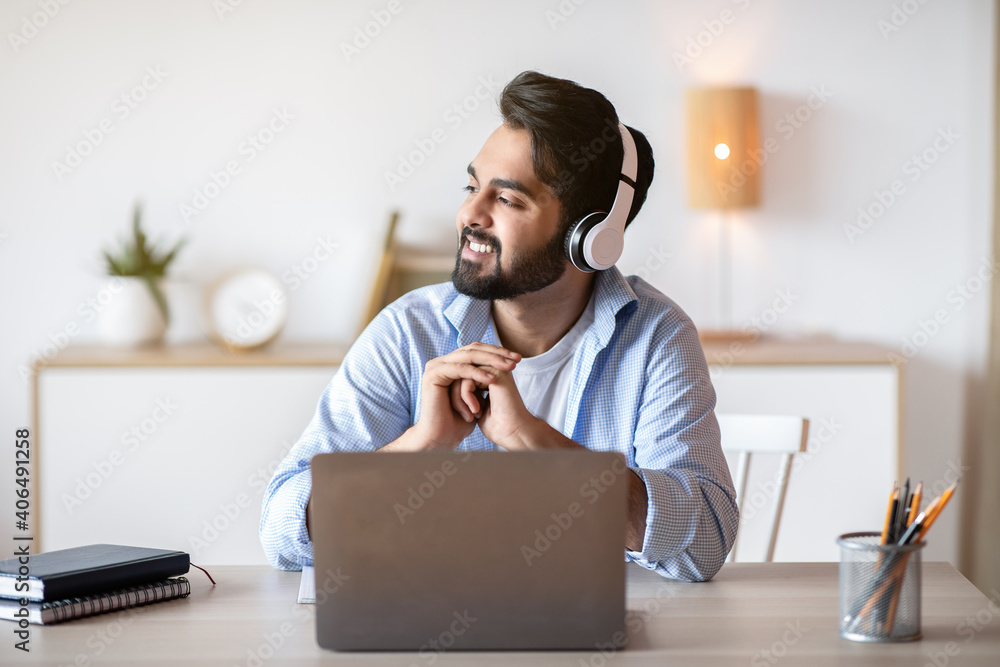 Smiling arab man in headphones listening music while working on laptop - obrazy, fototapety, plakaty 