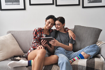 Wall Mural - Happy young lesbian couple hugging having fun using digital tablet relaxing on couch at home. Two smiling women friends holding computer looking at screen enjoying surfing online watching videos.