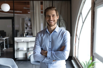 Head shot portrait smiling successful young businessman standing with arms crossed in modern office, happy executive boss startup founder looking at camera, posing for corporate photo alone