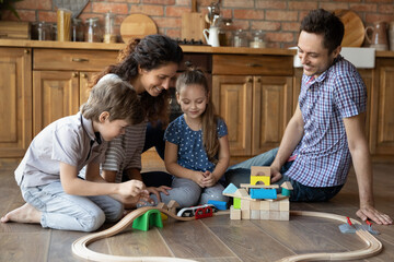 Wall Mural - Overjoyed young Caucasian family with small kids sit on floor in kitchen play toys together. Happy mom and dad have fun feel playful engaged in game activity relax with little children at home.