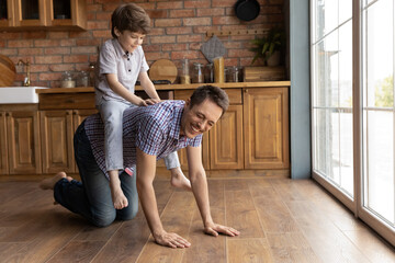 Wall Mural - Excited little 6s boy child ride happy young Caucasian father, engaged in funny game at home together. Overjoyed small kid sit on playful smiling dad back play in cozy kitchen on family weekend.