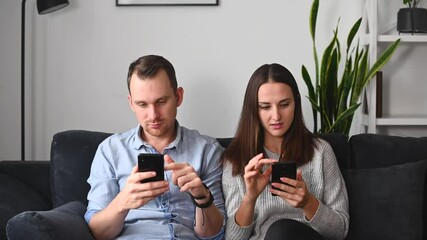 Couple sits on the comfortable couch and using mobile phones separately, a man and a woman lost in a network, do not pay attention to each other
