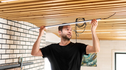 A male electrician is installing electrical wiring in the house. The rent-a-gent changes the lights on the stylish wooden planks ceiling. A builder is installing light into a designer ceiling.