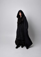 Wall Mural - Full length portrait of pretty black haired woman wearing long dark gown and a cloak.  Standing pose facing away from the camera, against a  studio background.