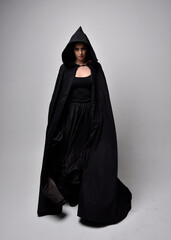 Sticker - Full length portrait of pretty black haired woman wearing long dark gown and a cloak.  Standing pose facing away from the camera, against a  studio background.