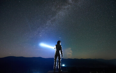 Cosmonaut wearing white space suit and helmet directs a blue ray of light into starry sky above horizon in the night, standing with his back to the camera in the mountains, space travel concept