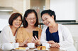 © DragonImages - Elderly Asian woman showing funny photos on smartpone screen to her friends when they are sitting at kitchen table