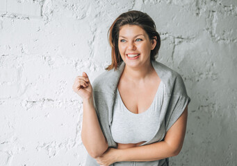 Beautiful happy emotional brunette young woman plus size body positive in comfortable sport wear near the white brick wall