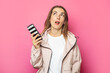 © Alex - Young surprised woman holding a phone, pink isolated background