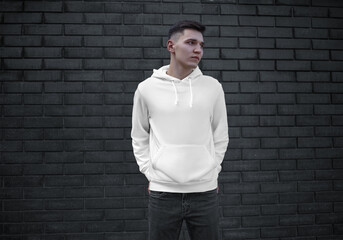 White hoodie template on a young guy with hands in pockets, blank mens clothing on a brick wall background, front view.