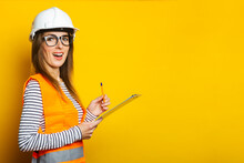 Young Woman With A Surprised Face In A Vest And Hard Hat Holds A Clipboard On A Yellow Background. Concept For Construction, New Building, Renovation. Banner